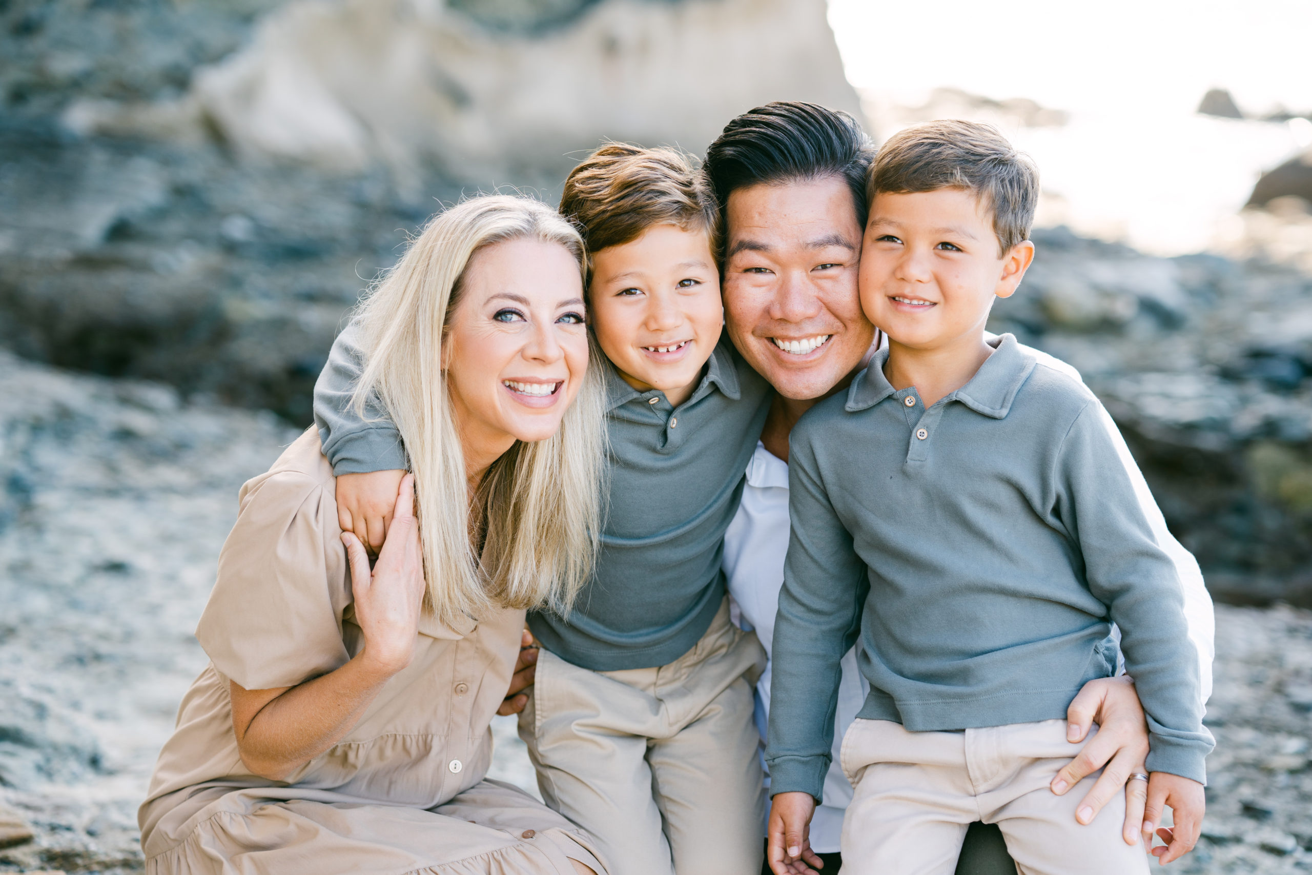 A family of a mom, a dad, and two boys smile together at Laguna Beach. Their heads are all pressed together.