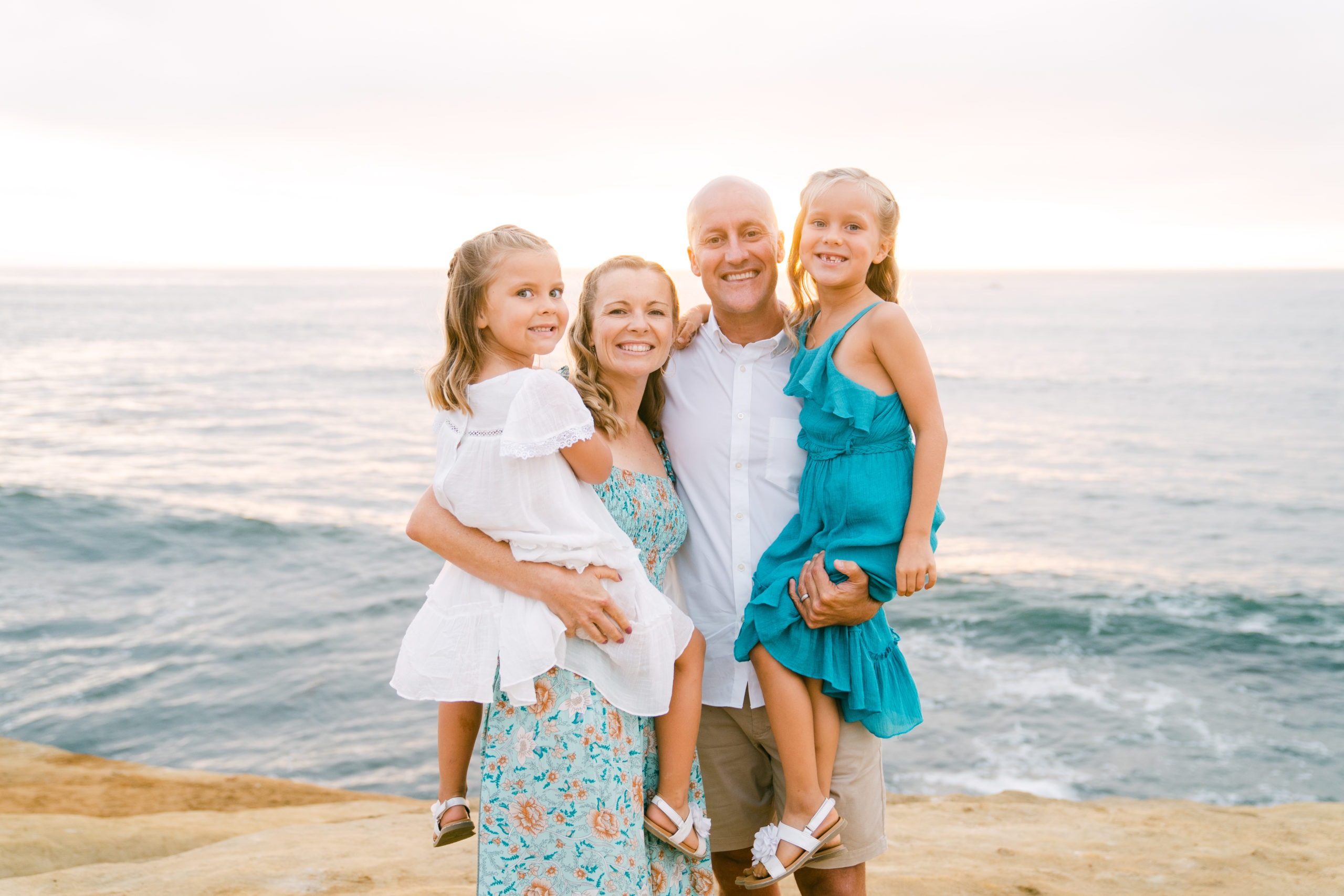 A white family smiles together at Sunset Cliffs Park in San Diego. The dad and mom are holding the two girls while the sun sets behind them over the water.