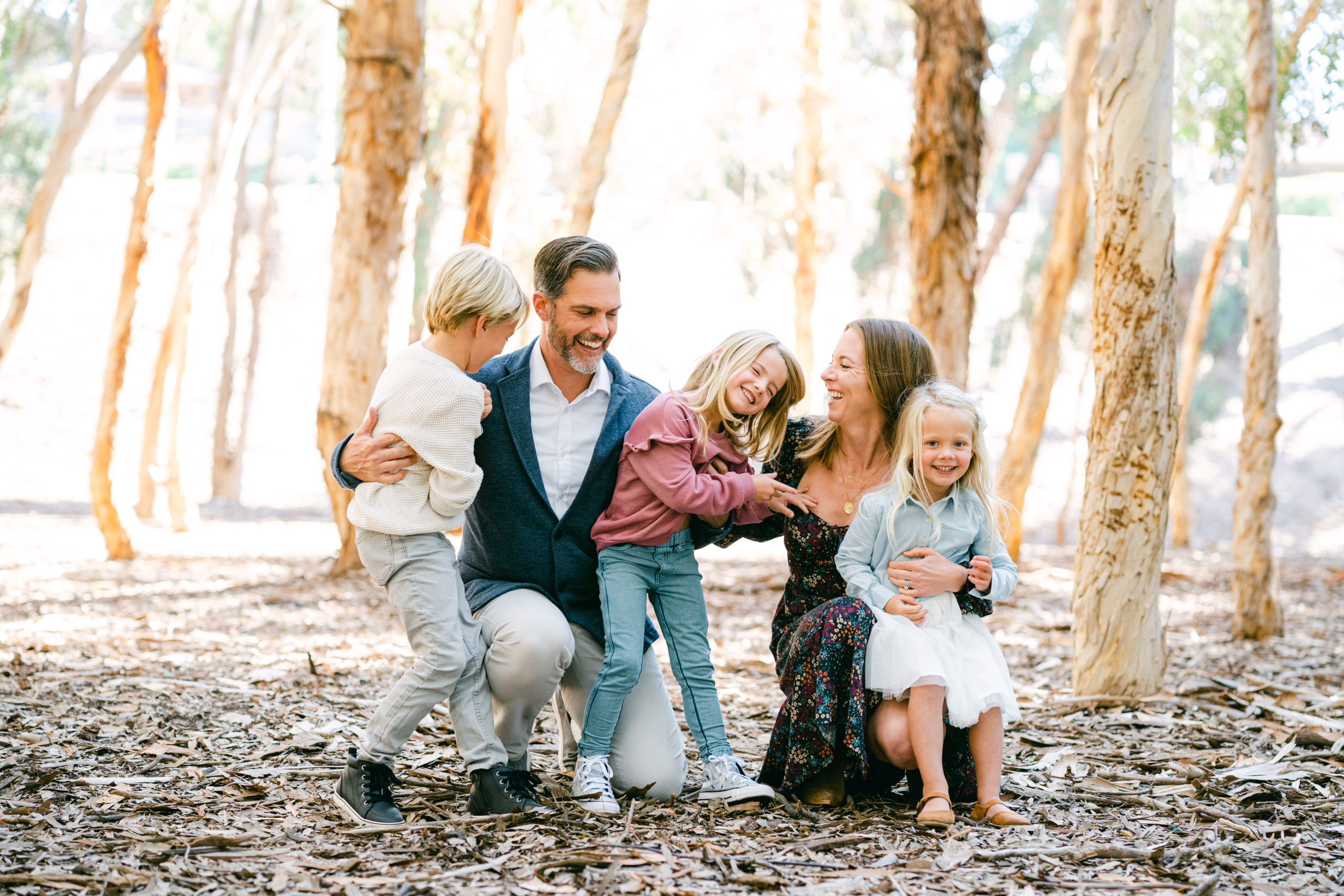 A White family of five laugh together at eucalyptus grove during an Orange County Mini Session. A dad, mom, two daughters, and a son.