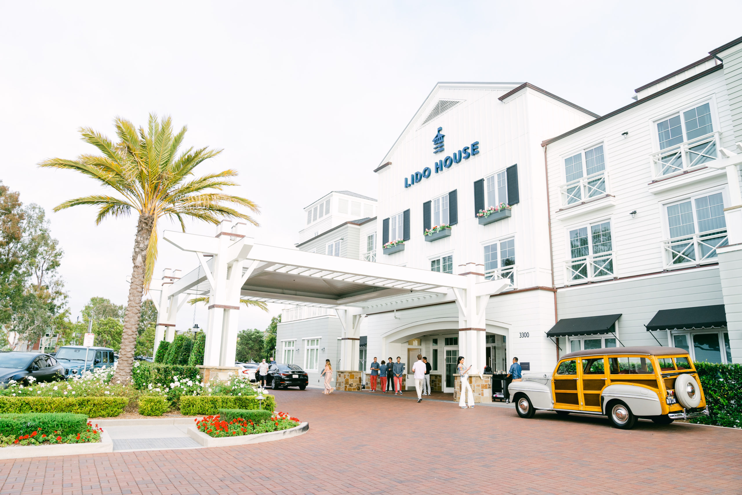 The Wedding Venue: The beautiful Lido House Hotel in Newport Beach. A white historic mansion with navy trim. Out front stands a lush Southern California landscape and a classic station wagon.