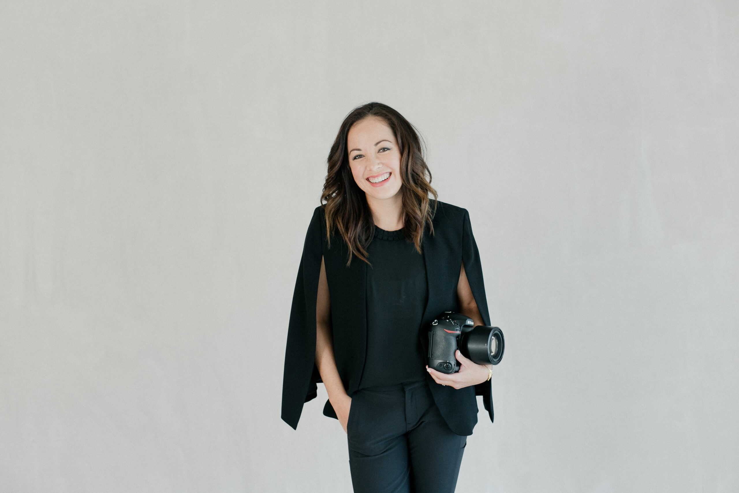 Kate stands wearing a black suit and smiles to camera as she holds her black camera by her side